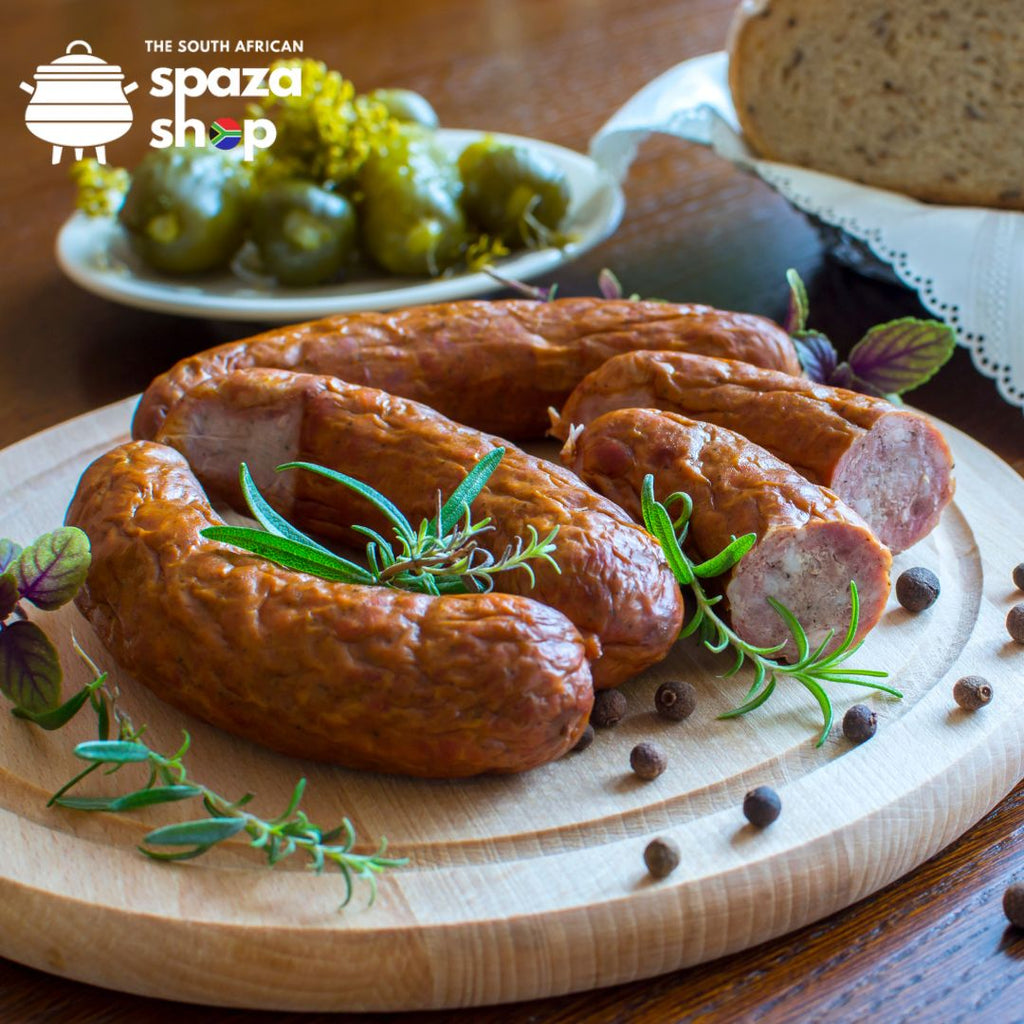 Traditional South African smoked sausages served on a wooden board with fresh rosemary, black peppercorns, rye bread, and pickled gherkins – authentic flavours available at The South African Spaza Shop in the UK.